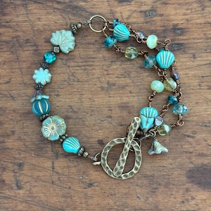 May include: A handcrafted bracelet featuring turquoise, teal, and green beads. The bracelet includes floral and striped beads, with a hammered bronze clasp. The design incorporates various shapes and textures, creating a unique accessory.