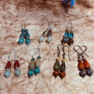 May include: A collection of handmade earrings with various designs and colors. The earrings feature beads in shades of blue, turquoise, red, orange, brown, and green. Some earrings have silver-colored hooks, while others have darker metal hooks.