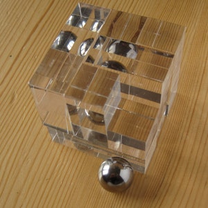 Puede incluir: Un cubo de vidrio transparente con múltiples compartimentos, cada uno con una bola de rodamiento de plata. Una sola bola de rodamiento de plata se encuentra fuera del cubo.