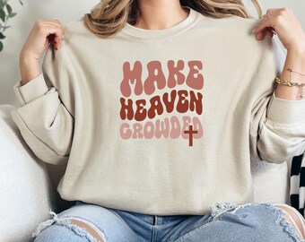 May include: A beige sweatshirt with the text "MAKE HEAVEN CROWDED" in a retro font with a red cross. The text is in shades of pink and brown.
