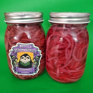 May include: Two glass jars filled with pickled red onions. The jars have silver lids and a label that reads "Kelly's Death Pickles" with a cartoon skull and crossbones graphic. The label also says "Atlanta, GA".