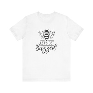 Könnte beinhalten: Weißes T-Shirt mit einer schwarz-weißen Bienenillustration und dem Text "LET'S GET BUZZED". Die Bienenillustration befindet sich über dem Text in einer kursiven Schriftart. Das T-Shirt hat kurze Ärmel und einen Rundhalsausschnitt.
