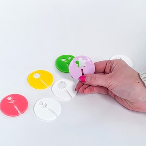 May include: A set of six colorful wine glass charms featuring different flower designs. The charms are made of acrylic and have a hole in the center for attaching to a wine glass stem. The charms are pink, green, yellow, and white.