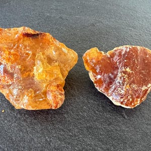 May include: Two raw, unpolished amber stones with irregular shapes. The larger stone on the left is a translucent golden yellow, while the smaller stone on the right is a deeper, semi-transparent reddish-brown with a smooth, glossy surface texture.