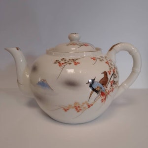 May include: White porcelain teapot with a bamboo handle, featuring a painted design of birds and a mountain landscape. The teapot has a lid with a knob on top.