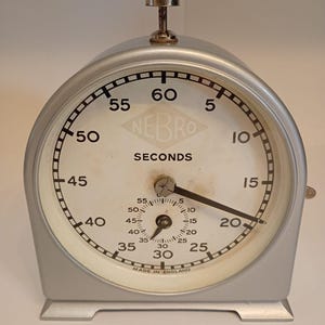 May include: A silver-coloured analogue stopwatch with a white face, featuring the brand name NEBRO, the word SECONDS, and the text MADE IN ENGLAND. The device has a large main dial, a smaller sub-dial, and a metallic winding knob on the top.