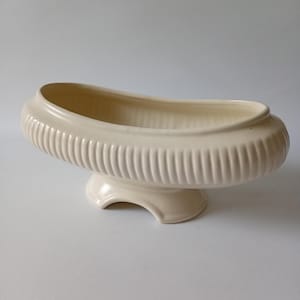 May include: An oval-shaped cream-coloured ceramic planter with a glossy finish. The exterior features a vertical ribbed texture around the perimeter. The vessel is supported by a pedestal base with a curved cutout design on the bottom.
