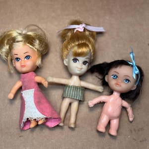 May include: Three vintage plastic dolls with blonde, light brown, and black hair. The dolls have blue eyes and are wearing a pink dress with a white apron, a green and white striped top, and a blue bow. The dolls are small and appear to be from the mid-20th century.