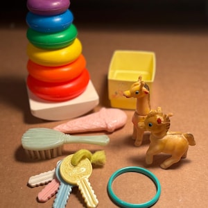 May include: A set of vintage children's toys including a colorful stacking ring toy, a yellow toy box, a giraffe, a horse, a brush, a set of keys, and a turquoise ring.