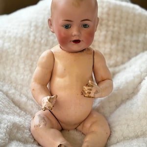 May include: A vintage porcelain doll with blue eyes and rosy cheeks, sitting on a white fluffy blanket. The doll is wearing no clothes and has a painted smile.