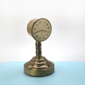 May include: A gold-colored clock with Roman numeral markings on a decorative stand. The clock face is white with black markings and hands.