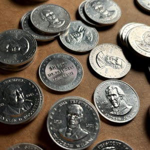 May include: A collection of silver-colored coins featuring different U.S. presidents. The coins are arranged in a random pattern on a brown surface. The coins feature the president's name, year of presidency, and the words "Mr. President" and "Coin Game".