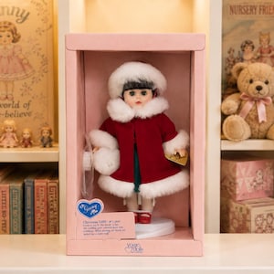 May include: A vintage Ginny doll in a pink box. The doll wears a red coat with white fur trim, a matching hat, and red shoes. The box has a heart-shaped tag that reads "Ginny" and "Christmas 1959".