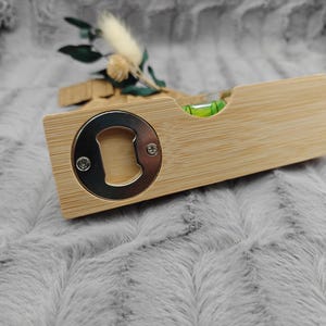 May include: A wooden bottle opener with a built-in level. The opener has a silver metal cap lifter and a small green level bubble. The wood has a light brown color. Decorative sprigs are in the background.