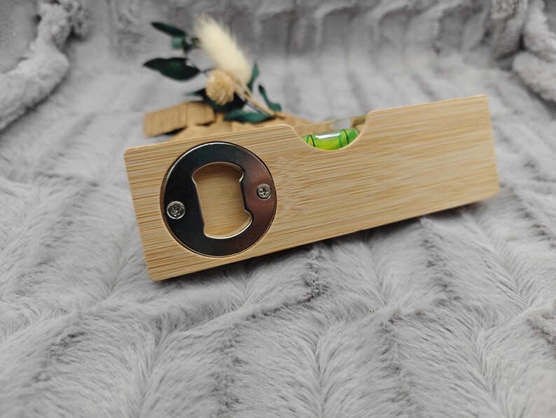 May include: A wooden bottle opener with a built-in level. The opener has a silver metal cap lifter and a small green level bubble. The wood has a light brown color. Decorative sprigs are in the background.