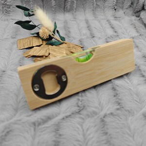 May include: A wooden bottle opener with a built-in level and a silver-colored opener. The opener is rectangular and made of light-colored wood. A small green bubble level is visible. Decorative dried flowers and wood pieces are in the background.