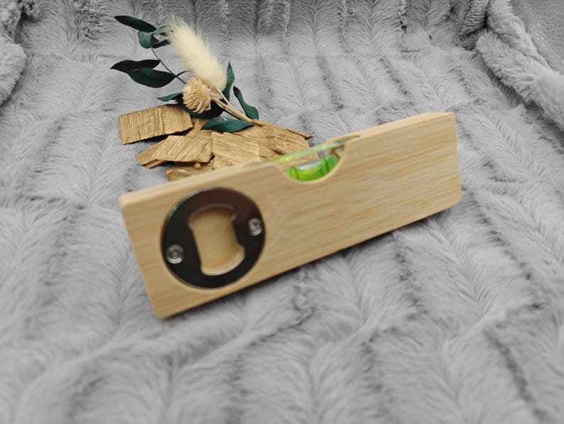 May include: A wooden bottle opener with a built-in level and a silver-colored opener. The opener is rectangular and made of light-colored wood. A small green bubble level is visible. Decorative dried flowers and wood pieces are in the background.