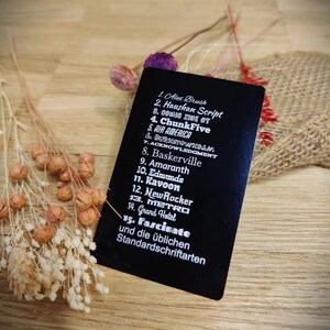 May include: A black rectangular card with a list of font names in white text. The card is angled on a wooden surface, with dried flowers and burlap in the background. The text includes "Alex Brush", "Haushan Script", and "Fascinate".