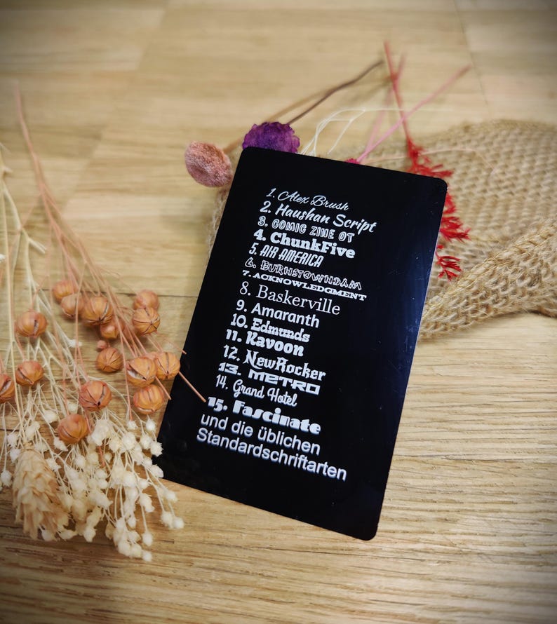 May include: A black rectangular card with a list of font names in white text. The card is angled on a wooden surface, with dried flowers and burlap in the background. The text includes "Alex Brush", "Haushan Script", and "Fascinate".