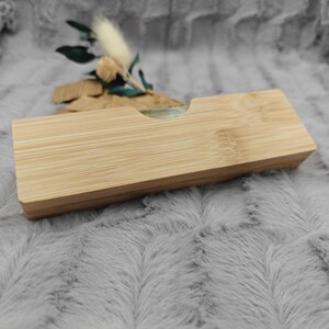 May include: A rectangular bamboo box with a recessed top. The box is made of light-colored wood with visible grain. A small arrangement of dried flowers and greenery sits behind the box. The background is a soft, gray, textured fabric.