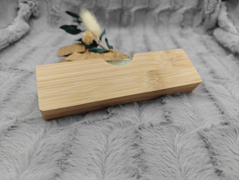 May include: A rectangular bamboo box with a recessed top. The box is made of light-colored wood with visible grain. A small arrangement of dried flowers and greenery sits behind the box. The background is a soft, gray, textured fabric.