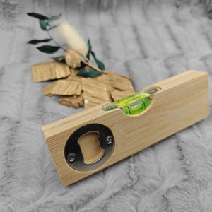 May include: A bamboo level and bottle opener combination. The level has a clear vial with a green liquid bubble. The bottle opener is a circular metal piece. The item is on a gray, textured surface.