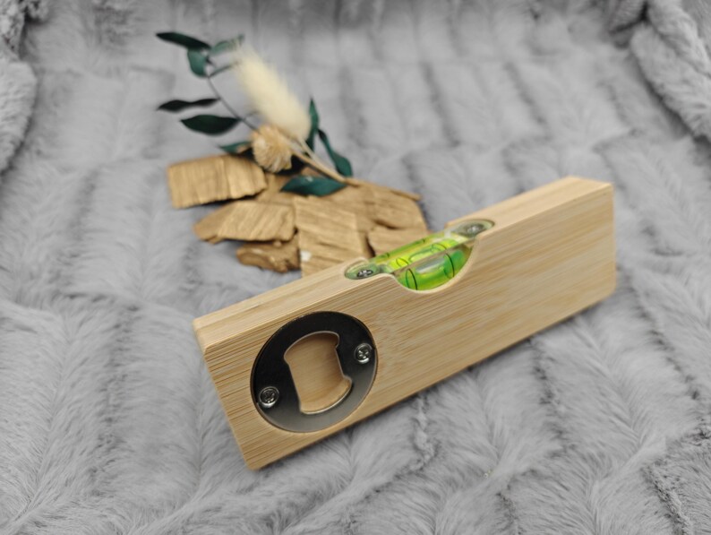 May include: A bamboo level and bottle opener combination. The level has a clear vial with a green liquid bubble. The bottle opener is a circular metal piece. The item is on a gray, textured surface.