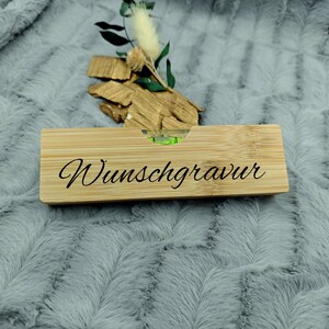 May include: A rectangular bamboo level with the word "Wunschgravur" in black script. The level has a small bubble indicator and is resting on a gray, textured surface. Decorative wood pieces and greenery are behind the level.