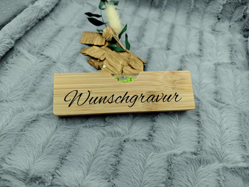 May include: A rectangular bamboo level with the word "Wunschgravur" in black script. The level has a small bubble indicator and is resting on a gray, textured surface. Decorative wood pieces and greenery are behind the level.