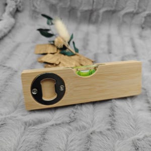 May include: A wooden bottle opener with a built-in level. The opener has a silver metal bottle cap remover on one end and a green bubble level in the center. The wood has a light brown color.