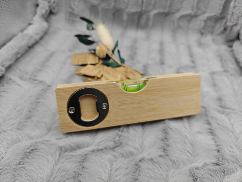May include: A wooden bottle opener with a built-in level. The opener has a silver metal bottle cap remover on one end and a green bubble level in the center. The wood has a light brown color.