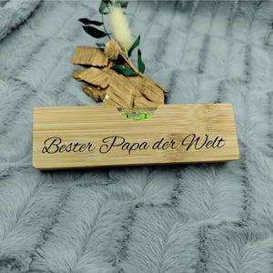 May include: A rectangular bamboo level with the text "Bester Papa der Welt" engraved on it. The level has a green bubble indicator. The item is decorated with small pieces of wood and greenery.
