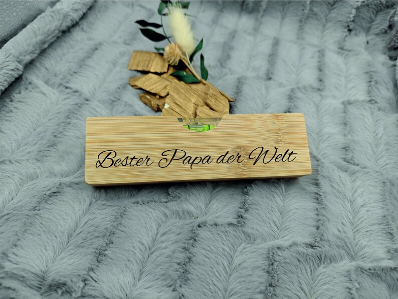 May include: A rectangular bamboo level with the text "Bester Papa der Welt" engraved on it. The level has a green bubble indicator. The item is decorated with small pieces of wood and greenery.