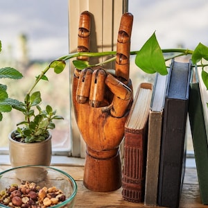 May include: A wooden articulated hand sculpture posed in a rock and roll gesture. The hand features a warm brown finish with visible wood grain, a cylindrical base, and jointed fingers positioned with the index and pinky fingers extended upward.