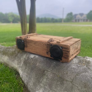 May include: A wooden box with two black speaker grills and metal hardware. The box is made of weathered wood and sits on a stone surface. The background features a blurred green field and trees.