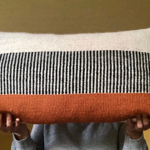 May include: Rectangular decorative pillow with a woven design. The pillow features three horizontal bands: a top white section, a middle section with black and white vertical stripes, and a bottom rust-colored section. The edges have a fringed trim.
