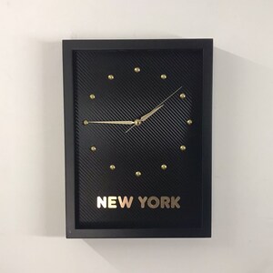 May include: Black framed clock with gold accents and a carbon fibre face. The clock face has gold hour markers and the words "NEW YORK" are printed in gold letters below the clock face.