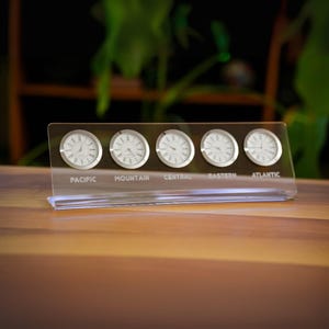 May include: A clear acrylic desk clock displaying five time zones: Pacific, Mountain, Central, Eastern, and Atlantic. Each clock face is white with black hands and silver trim. The base is clear with the time zone names printed in white.