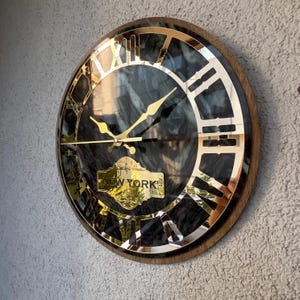 Può includere: Orologio da parete rotondo con quadrante effetto marmo scuro, numeri romani dorati e lancette dorate. L'orologio ha una cornice in legno e una placca dorata con la scritta "NEW YORK".