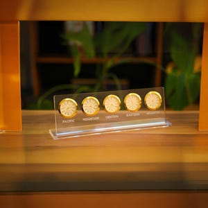 May include: A clear acrylic desk clock displaying five small clocks with gold-colored frames. Each clock face shows a different time zone: Pacific, Mountain, Central, Eastern, and Atlantic. The clock sits on a wooden surface.
