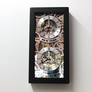 May include: Black framed clock with two silver clock faces. The top clock face has the words "New York" and the bottom clock face has the words "London".