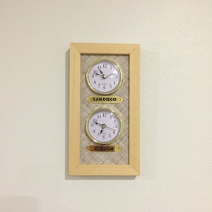 May include: A wooden framed clock with two analog clock faces. The top clock face reads "SAN DIEGO" and the bottom clock face reads "LONDON".