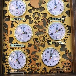 May include: A wall clock with six gold-colored clock faces, each with a different time zone. The time zones are Pacific, Mountain, Eastern, Central, Hawaii, and London. The clock faces are surrounded by gold-colored gears.