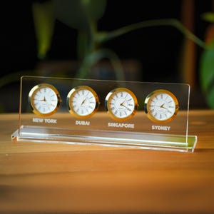 May include: A clear acrylic desk clock displaying four analog clocks with gold-colored bezels. Each clock face is white with black Roman numerals and hands. The clock displays the time for New York, Dubai, Singapore, and Sydney.