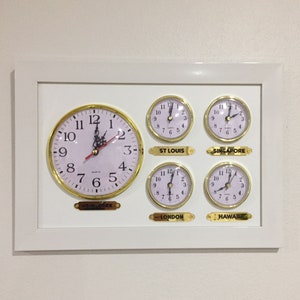 Può includere: Un orologio da parete incorniciato in bianco con cinque orologi più piccoli all'interno. Gli orologi mostrano l'ora a New York, St. Louis, Singapore, Londra e Hawaii.