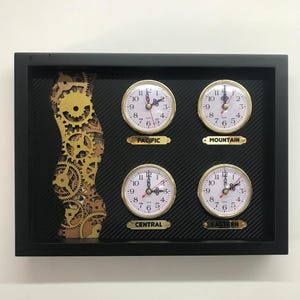 May include: A black framed wall clock with four gold-rimmed analog clocks displaying different time zones. The clocks are labeled "Pacific", "Mountain", "Central", and "Eastern".