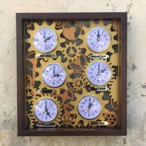 May include: A wooden wall clock with six gold-coloured clock faces, each displaying a different time zone: Pacific, Mountain, Central, Eastern, Hawaii, and London. The clock faces are surrounded by gold-coloured gears.
