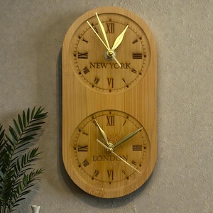 May include: A wooden wall clock with two circular faces, one above the other. The top clock face reads "NEW YORK" and the bottom reads "LONDON". Both faces have gold hands and Roman numeral hour markers. The clock is made of light-colored wood.