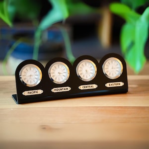 May include: A black desk clock with four clock faces, each displaying a different time zone: Pacific, Mountain, Central, and Eastern. The clock has a rectangular base and a curved top. The clock faces have white backgrounds with black Roman numerals and hands.