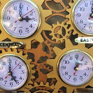 May include: A gold-colored wall clock with four round analog clocks, each with a different time zone: Central, Eastern, London, and a fourth time zone. The clocks are connected by gears and have a gold-colored background.